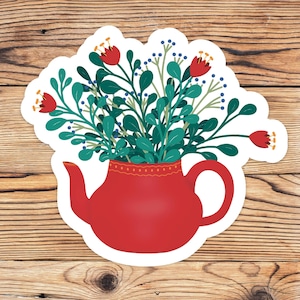 Op de afbeelding: Een rode theepot met een witte en rode gepatroneerde rand gevuld met een boeket groene bladeren en rode bloemen.