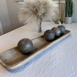 May include: A wooden serving tray with a rustic finish, featuring four dark gray round balls arranged in a row. The tray is on a white textured surface.