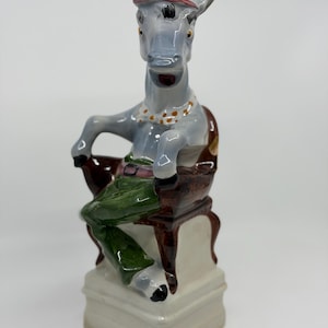 Peut inclure: Une figurine en céramique représentant un âne gris assis sur une chaise marron. L'âne porte un haut-de-forme rayé rouge, blanc et bleu, un nœud papillon blanc à pois orange et un pantalon vert avec une ceinture rose sur un socle rectangulaire.