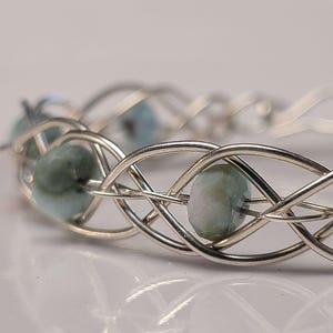 May include: A silver-toned bracelet with a woven design, featuring light blue-green gemstone beads. The bracelet's intricate metalwork creates a delicate, intertwined pattern, showcasing the gemstones' subtle color and faceted surfaces.
