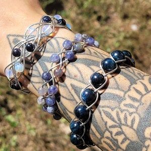 May include: Three wire-wrapped bracelets with different colored gemstones. The top bracelet has iridescent opal stones, the middle bracelet has blue and gray stones, and the bottom bracelet has dark blue tiger's eye stones.