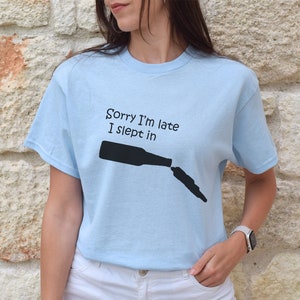 May include: A light blue t-shirt with a black graphic of a beer bottle and the text "Sorry I'm late I slept in".