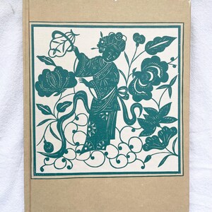 May include: A vintage book with a tan cover and a teal illustration. The illustration features a woman in traditional attire surrounded by flowers and foliage. The artwork is framed within a teal border.