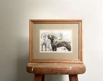 May include: A framed print featuring three dogs in a black and white illustration. The artwork is set within a light brown wooden frame and a green mat. The frame is displayed on a rustic wooden stool.