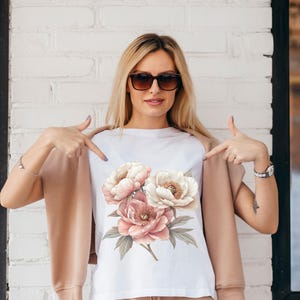 May include: A white t-shirt with a floral print of pink and white peonies. The model is wearing the shirt and pointing to it with both hands.