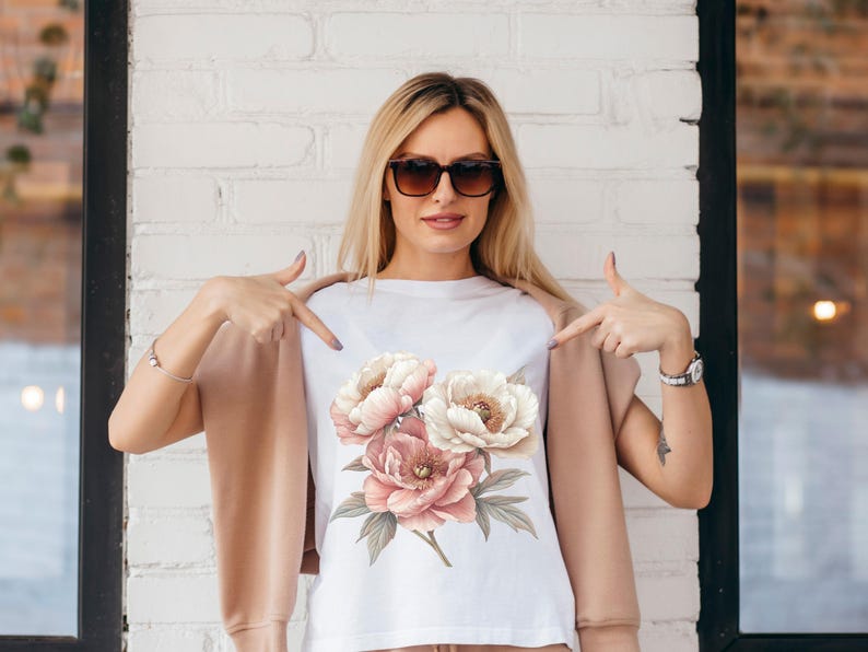 May include: A white t-shirt with a floral print of pink and white peonies. The model is wearing the shirt and pointing to it with both hands.