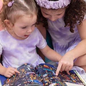 May include: Two young girls in purple tie-dye shirts are looking at a book with colorful illustrations. The book is open and the girls are pointing at the pictures.