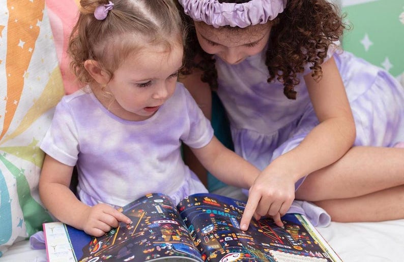 May include: Two young girls in purple tie-dye shirts are looking at a book with colorful illustrations. The book is open and the girls are pointing at the pictures.