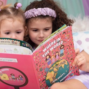 May include: Two young girls are reading books. The girl on the left is reading a book titled "Characterful" with a magnifying glass on the cover. The girl on the right is reading a book titled "Where in the World Are Brett, Maddison & Made 2?" with a colorful world map on the cover.
