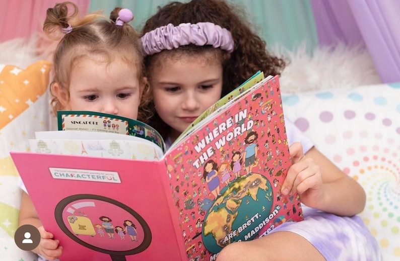 May include: Two young girls are reading books. The girl on the left is reading a book titled "Characterful" with a magnifying glass on the cover. The girl on the right is reading a book titled "Where in the World Are Brett, Maddison & Made 2?" with a colorful world map on the cover.