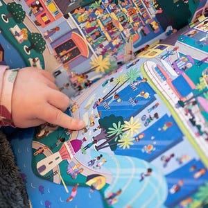 May include: A colorful children's book with a busy city scene on one page and a beach scene on the other. The book is open and a child's hand is pointing at the beach scene.