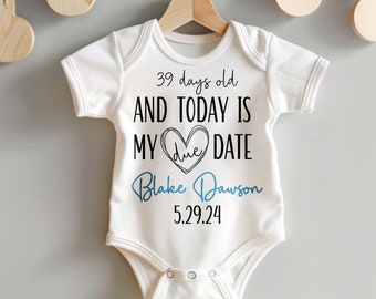 Puede incluir: Un body blanco para bebé con texto negro que dice "39 days old AND TODAY IS MY due DATE Blake Dawson 5.29.24". El texto "due" está dentro de un corazón.