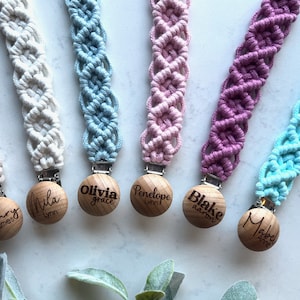 May include: Six colorful crocheted baby teethers with wooden clips and personalized engraved names. The teethers are in shades of white, blue, pink, purple, and turquoise. The names on the clips are Harry James, Mila Lynn, Olivia Grace, Penelope Lynn, Blake Aaron, and Mabel Anne.
