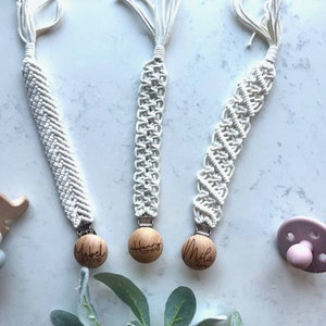 May include: Three white crocheted pacifier clips with wooden beads. The beads are engraved with the names "Mabel Anne", "Henry", and "Mila".