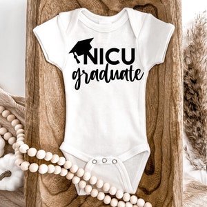 May include: White baby onesie with black text that reads "NICU graduate" and a black graduation cap graphic.