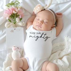May include: A white baby onesie with the text "tiny but mighty" in black lettering. A baby is wearing the onesie and sleeping on a white blanket. The baby is wearing a headband with a flower on it.