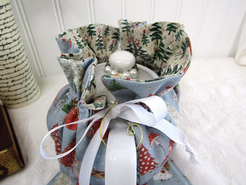 May include: A blue teapot cozy with a white ribbon and a Christmas tree print. The cozy is lined with a beige fabric with a green Christmas tree print.