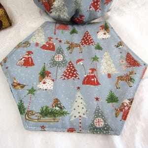 May include: A blue hexagonal fabric placemat with a Christmas theme. The placemat features a repeating pattern of red and green Christmas trees, reindeer, and people wearing red and white clothing. The placemat is edged with a light blue stitching.