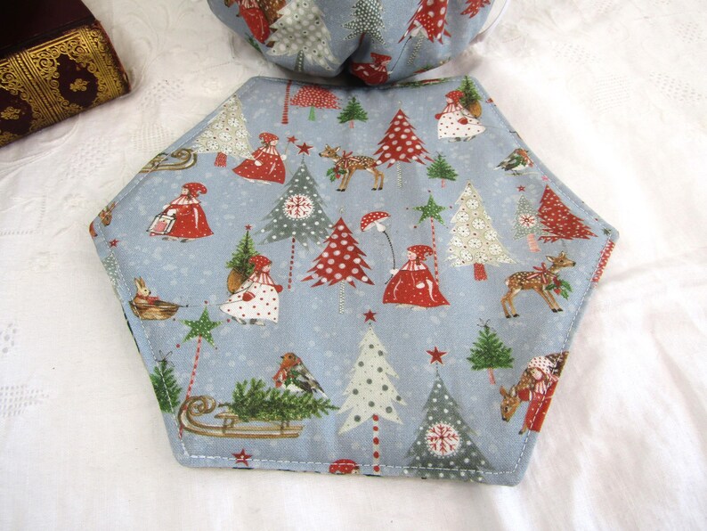 May include: A blue hexagonal fabric placemat with a Christmas theme. The placemat features a repeating pattern of red and green Christmas trees, reindeer, and people wearing red and white clothing. The placemat is edged with a light blue stitching.