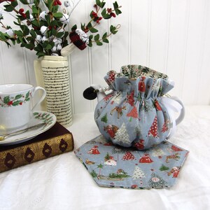 May include: A blue teapot cozy with a Christmas tree pattern. The cozy is decorated with red, white, and green accents. The teapot cozy is sitting on a blue and white patterned tea cozy.