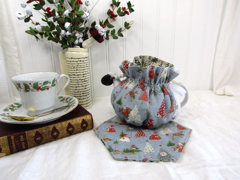 May include: A blue teapot cozy with a Christmas tree pattern. The cozy is decorated with red, white, and green accents. The teapot cozy is sitting on a blue and white patterned tea cozy.