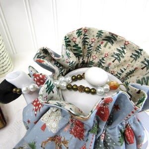 May include: A blue teapot cozy with a white background and a Christmas tree pattern. The cozy is lined with a cream fabric with a green Christmas tree pattern. The teapot cozy is decorated with a white pearl necklace and a brown beaded necklace.