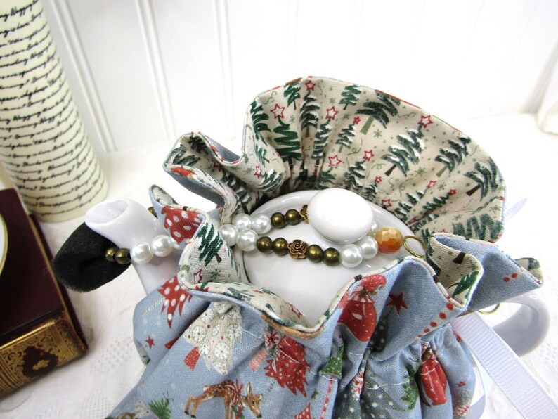 May include: A blue teapot cozy with a white background and a Christmas tree pattern. The cozy is lined with a cream fabric with a green Christmas tree pattern. The teapot cozy is decorated with a white pearl necklace and a brown beaded necklace.