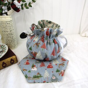 May include: A blue and white teapot cozy with a Christmas tree pattern. The cozy is made of fabric and has a drawstring closure. The cozy is sitting on a blue and white fabric coaster with a Christmas tree pattern.
