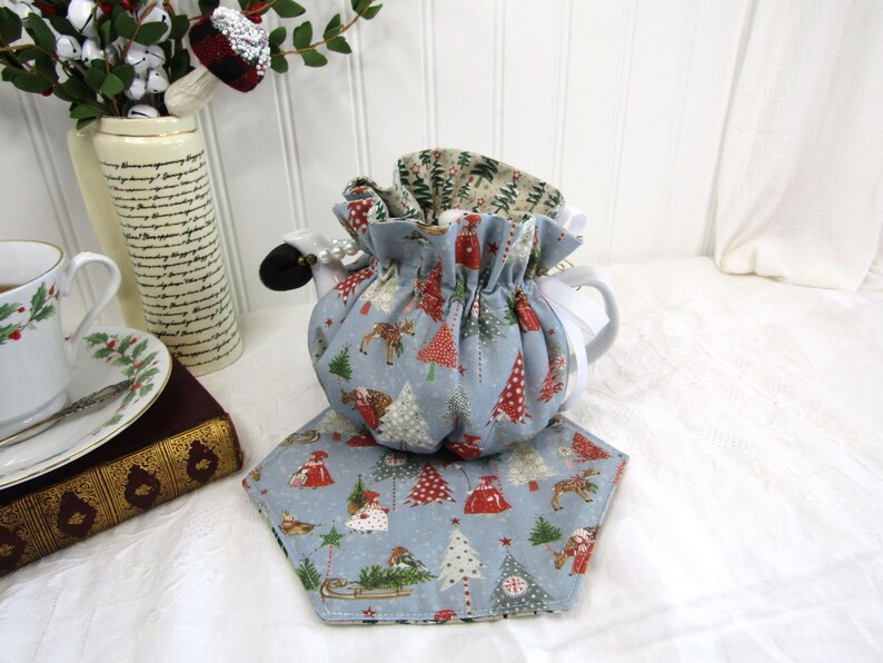 May include: A blue and white teapot cozy with a Christmas tree pattern. The cozy is made of fabric and has a drawstring closure. The cozy is sitting on a blue and white fabric coaster with a Christmas tree pattern.