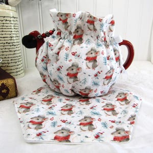 Può includere: Copertura per teiera bianca con stampa di topo rosso e bianco. La copertura è raccolta in alto e ha un nastro rosso. La teiera è rossa e ha un manico. La copertura è appoggiata su un sottobicchiere esagonale bianco con la stessa stampa di topo.