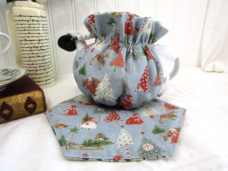 May include: A blue teapot cozy with a Christmas tree and reindeer print. The cozy has a white drawstring and is sitting on a matching blue and white Christmas tree print placemat.