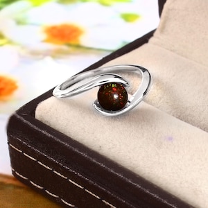 Könnte beinhalten: Ein silberner Ring mit einem dunklen, runden Edelstein mit roten und grünen Reflexen. Das Design des Rings ist fließend und offen, umschließend den Stein. Der Ring wird in einer braunen Schmuckschatulle mit cremefarbenem Innenraum präsentiert.