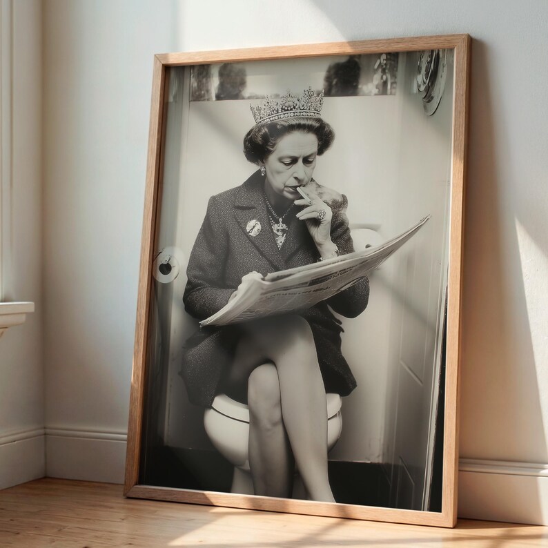 May include: Black and white framed print of a person wearing a crown and jewelry, seated on a toilet, smoking, and reading a newspaper. The print is in a light-colored wooden frame, leaning against a wall.