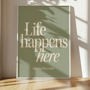 Peut inclure: Impression d'art encadrée avec l'inscription "Life happens here" en beige clair sur fond vert. La phrase "Let's stay for a while" est imprimée en dessous. L'impression est exposée sur un sol en bois.