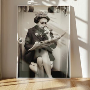 May include: Black and white framed print of a person wearing a crown, jewelry, and a blazer, sitting on a toilet, smoking a cigarette, and reading a newspaper. The print is in a white frame.