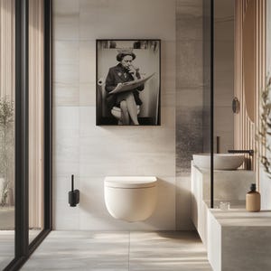 May include: A modern bathroom featuring a wall-mounted toilet with a soft, curved design. A framed black and white photograph hangs above the toilet. The bathroom has a minimalist aesthetic with neutral tones and a large window.
