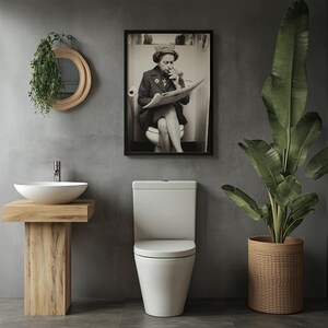 May include: A modern bathroom scene featuring a white toilet, a wooden vanity with a white sink, and a framed black and white print of a person reading a newspaper. A large potted plant in a woven basket adds a natural element to the space.