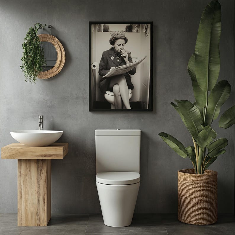 May include: A modern bathroom scene featuring a white toilet, a wooden vanity with a white sink, and a framed black and white print of a person reading a newspaper. A large potted plant in a woven basket adds a natural element to the space.