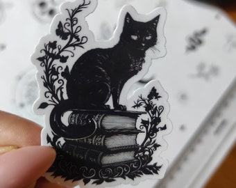 May include: A black cat sticker perched atop a stack of books, with floral and butterfly accents. The sticker has a white border and is set against a background of patterned paper. A small orange pumpkin is visible in the background.