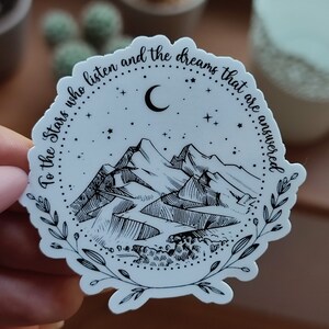 May include: A white sticker with a black and white illustration of mountains, a crescent moon, and stars. The text around the image reads "To the stars who listen and the dreams that are answered."