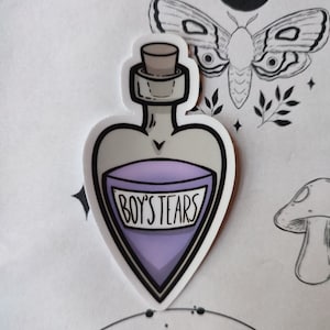 Könnte beinhalten: Ein herzförmiger Aufkleber mit einer lilafarbigen Zaubertrankflasche, auf der in weißen Buchstaben "BOY'S TEARS" steht. Die Flasche hat einen Korkverschluss und ist mit schwarzem Rand versehen.