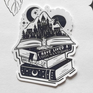 Puede incluir: Pegatina en blanco y negro que representa una pila de libros con una escena de castillo y bosque dentro del libro abierto. El texto "I have lived a thousand lives" está escrito en los libros.