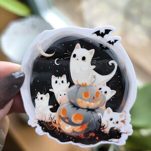 Könnte beinhalten: Ein weißer Aufkleber mit schwarzem Hintergrund, auf dem eine Gruppe weißer Katzen mit schwarzen Augen und Nasen zu sehen ist, die auf einem Stapel orangefarbener und grauer Jack-o'-Lantern-Kürbisse sitzen. Der Aufkleber hat einen Halbmond und Sterne im Hintergrund.