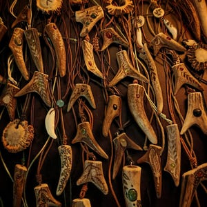 Pode incluir: Uma coleção de pingentes de chifre pendurados em finos cordões de couro marrom. Cada pingente apresenta padrões esculpidos, desenhos gravados e pedras preciosas coloridas incrustadas. Algumas peças incluem fatias circulares de chifre com runas.