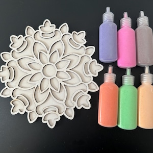 May include: A wooden mandala with a floral design, surrounded by six bottles of colorful sand art. The bottles have clear caps and are filled with purple, pink, brown, orange, green, and peach sand.