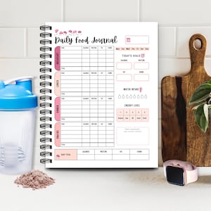 May include: A spiral-bound "Daily Food Journal" notebook with pink accents, designed for tracking meals, calories, protein, fat, and carbs. Includes sections for breakfast, lunch, dinner, and snacks, with a water intake tracker and energy level chart. A pink smartwatch and a protein shake are also visible.