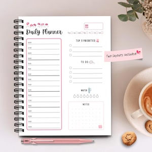 Può includere: Un'agenda giornaliera a spirale con un design rosa e bianco. L'agenda include sezioni per fasce orarie, priorità principali, liste di cose da fare, assunzione di acqua e note. Il testo "Daily Planner" è in alto. Due layout inclusi.