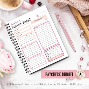 Puede incluir: Un planificador "Paycheck Budget" con espiral y detalles en rosa. El planificador incluye secciones para ingresos, gastos y seguimiento del presupuesto. Un bolígrafo rosa descansa junto al planificador abierto. El texto "Paycheck Budget" se muestra.