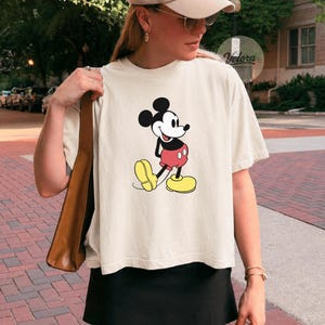 May include: Ivory-colored t-shirt featuring a classic Mickey Mouse graphic. The cartoon character is depicted in his iconic red shorts, yellow shoes, and black gloves. The shirt is paired with a black skirt.