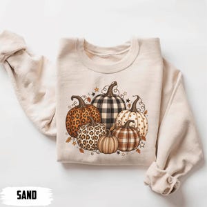 Puede incluir: Una sudadera de cuello redondo color arena con un gráfico central de cinco calabazas con varios patrones, incluyendo estampado de leopardo, cuadros vichy en blanco y negro y cuadros marrones, decorado con pequeñas estrellas y hojas de otoño.